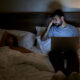 Hombre sentado en la oscuridad, sobre una cama y mirando una computadora. Mucha gente se pregunta si es malo ver pornografía. Te contamos por qué es malo ver porno solo en ciertas circunstancias.